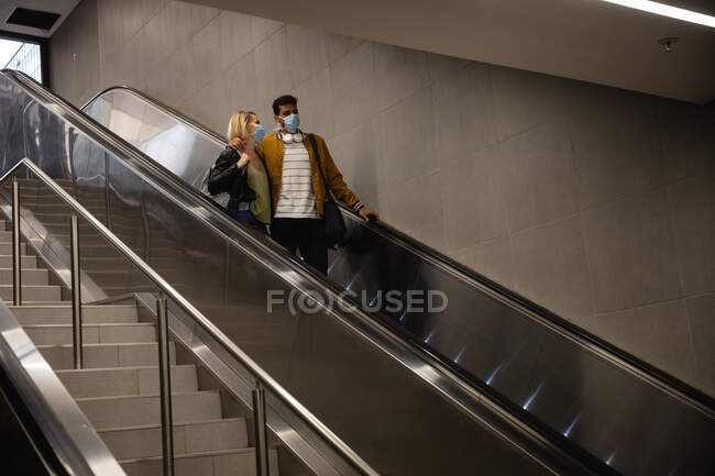 Vista frontal de ángulo bajo de una pareja caucásica en la ciudad, descendiendo en la estación subterránea con una escalera mecánica, usando máscaras faciales contra la contaminación del aire y covid19 coronavirus. - foto de stock