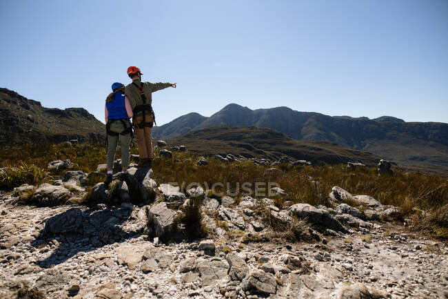 Vue arrière du couple caucasien profitant du temps dans la nature ensemble, portant un équipement de tyrolienne, debout admirant la vue, l'homme pointant sur une journée ensoleillée dans les montagnes — Photo de stock