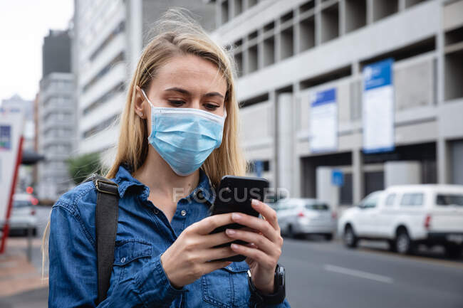 Nahaufnahme einer kaukasischen Frau mit Mundschutz gegen Luftverschmutzung und Covid19 Coronavirus, die mit ihrem Smartphone durch die Straßen der Stadt läuft. — Stockfoto