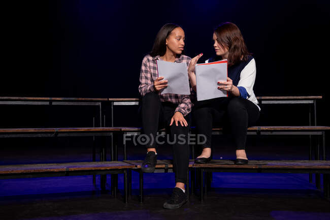 Vue de face d'une adolescente caucasienne et d'une adolescente métisse tenant des scénarios assis sur la scène d'un théâtre scolaire pendant les répétitions pour une représentation — Photo de stock