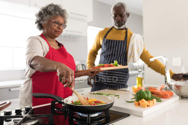Старшая афроамериканская пара проводит время дома вместе, социальное дистанцирование и самоизоляция в карантинной изоляции во время эпидемии коронавируса 19, стоя на кухне, готовясь к еде — стоковое фото
