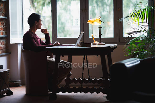 Autoaislamiento en cuarentena de encierro. vista lateral de un joven hombre de raza mixta, sentado en su oficina en casa, usando su computadora portátil mientras trabajaba. - foto de stock