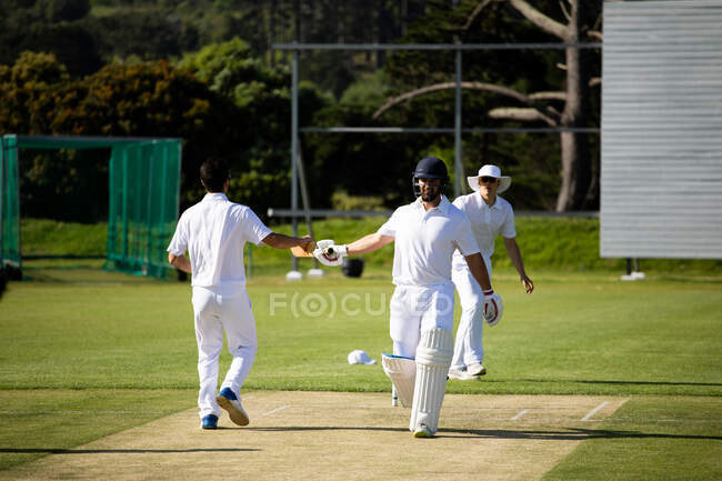 Vista frontal de dois jogadores de críquete adolescentes multi-étnicos que caminham pelo campo durante uma partida de críquete, mãos levantadas e punho-batendo, com outro jogador em pé no fundo. — Fotografia de Stock