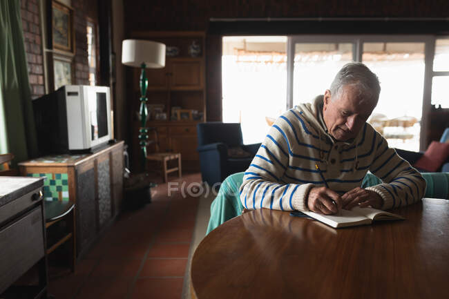 Vista lateral de um homem caucasiano sênior relaxando em casa, sentado à mesa em sua sala de jantar escrevendo em um livro com um lápis — Fotografia de Stock