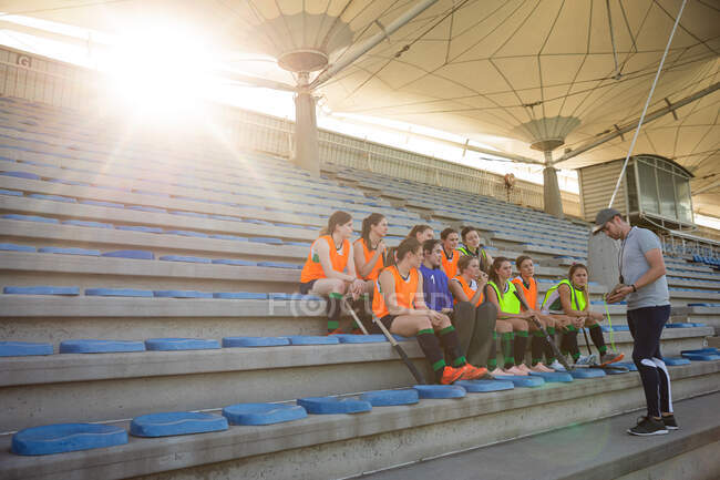 Вид сбоку на группу кавказских хоккеистов, готовящихся к матчу, сидящих на трибуне, держащих хоккейные клюшки, с их кавказским тренером по хоккею на траве, стоящим перед ними, говорящих о плане игры в солнечный день — стоковое фото