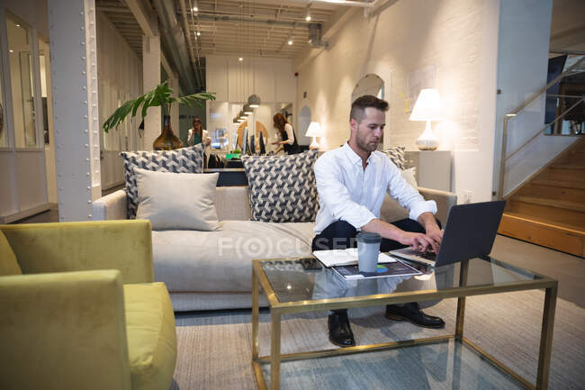Hombre caucásico negocio creativo trabajando en una oficina moderna informal, sentado en un sofá y el uso de un ordenador portátil con colegas que trabajan en el fondo - foto de stock