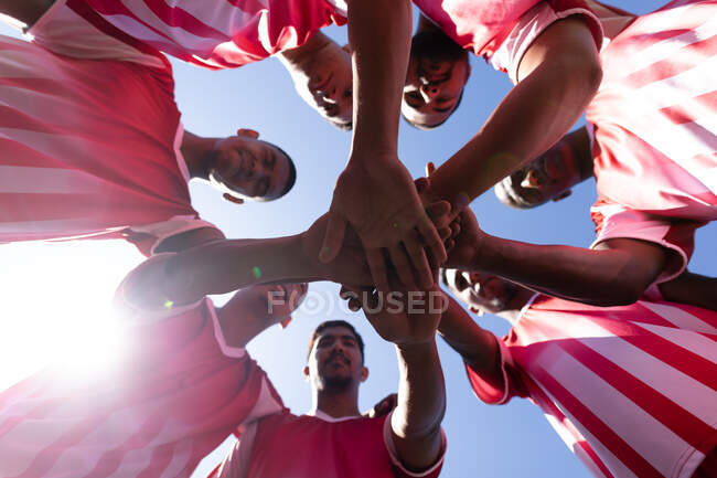 Tiefer Blickwinkel auf eine multiethnische Mannschaft von fünf Fußballern pro Seite, die auf einem Sportplatz in der Sonne ein Strip-Training tragen und im Kreis stehen und sich gegenseitig motivieren.. — Stockfoto