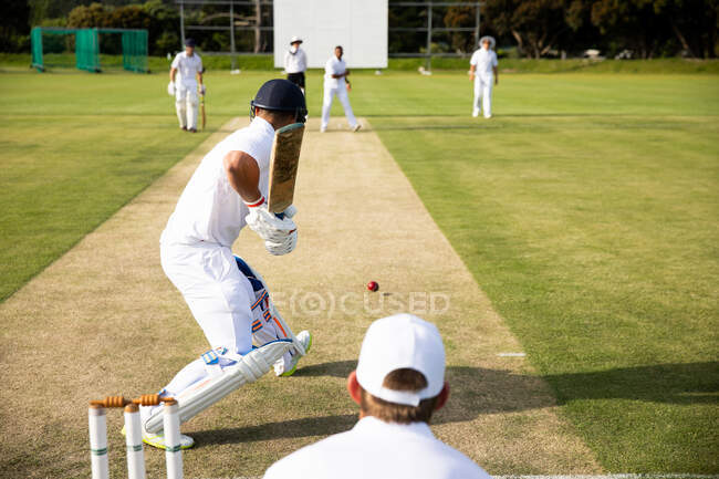 Visão traseira de um jogador de críquete masculino caucasiano adolescente em campo durante uma partida de críquete, segurando um taco de críquete pronto para bater uma bola de críquete com outros jogadores jogando atrás. — Fotografia de Stock