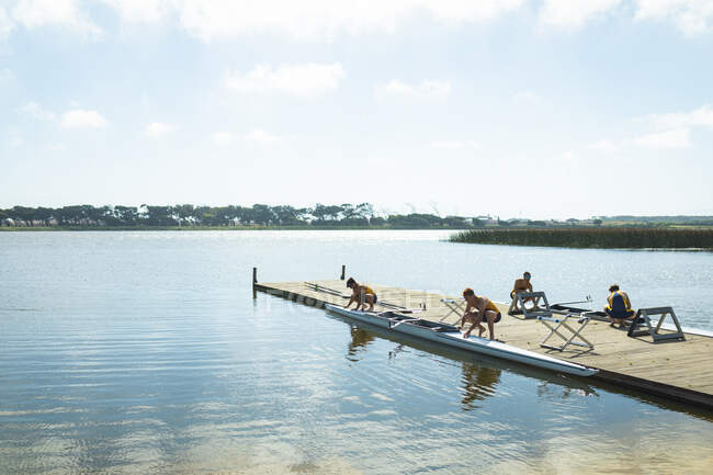 Vista lateral de uma equipe de remo de quatro homens caucasianos colocando barcos a remo na água antes de remar, de pé em um molhe no rio em um dia ensolarado — Fotografia de Stock