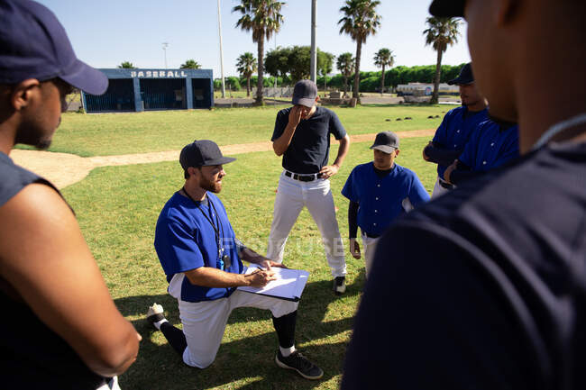 Vista lateral de un entrenador de béisbol masculino caucásico instruyendo a un equipo multiétnico de jugadores de béisbol masculinos, arrodillados, preparándolos antes de un juego en un campo de juego, en un día soleado - foto de stock