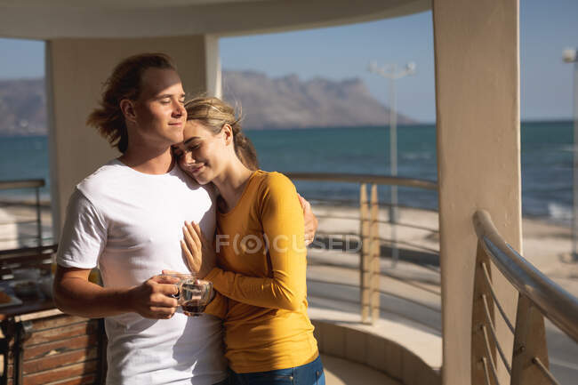 Kaukasisches Paar, das auf einem Balkon steht, sich umarmt und eine Tasse Kaffee hält. Soziale Distanzierung und Selbstisolierung in Quarantäne. — Stockfoto
