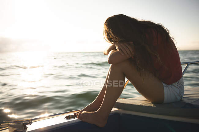 Una adolescente caucásica disfrutando de su tiempo de vacaciones en el sol junto a la costa, sentada en un barco, relajándose, cubriéndose la cara - foto de stock