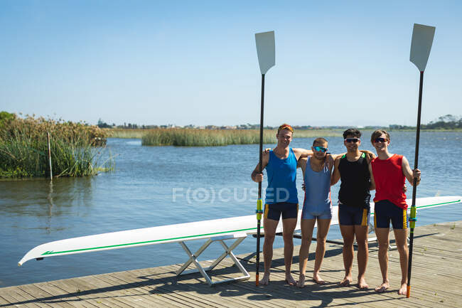 Front view of four Caucasian male rowers standing barefoot on a jetty holding oars and looking to camera, with a rowing boat in the background — Stock Photo