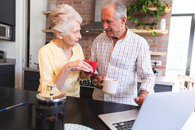 Una coppia di anziani caucasici in pensione a casa in piedi a un tavolo nella loro cucina, parlando e sorridendo, utilizzando un computer portatile e bevendo caffè insieme, coppia isolata durante la pandemia coronavirus19 — Foto stock