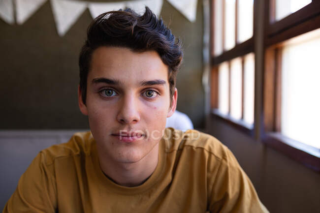 Portrait close up of a Caucasian teenage boy with dark hair and grey eyes sitting at a desk in a school classroom looking straight to camera — Stock Photo