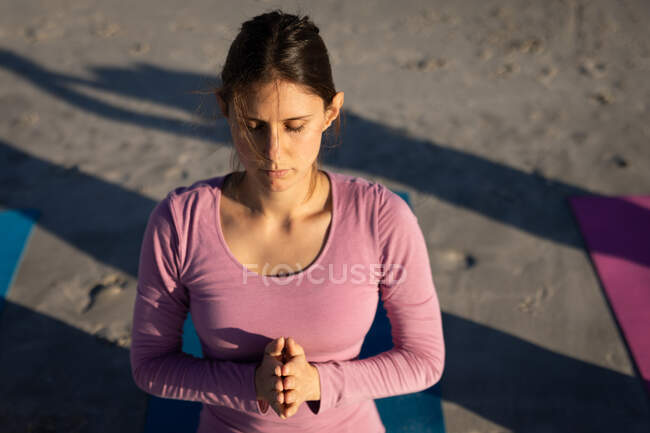 Angle élevé d'une femme caucasienne portant une chemise rose sur une plage ensoleillée, méditant, pratiquant le yoga les yeux fermés et les mains en position de prière. — Photo de stock