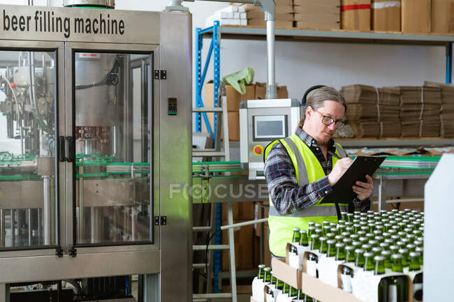 Homem caucasiano vestindo colete de alta visibilidade, trabalhando em uma microcervejaria, segurando um arquivo e escrevendo dados enquanto verifica garrafas de cerveja preparadas para entrega.. — Fotografia de Stock