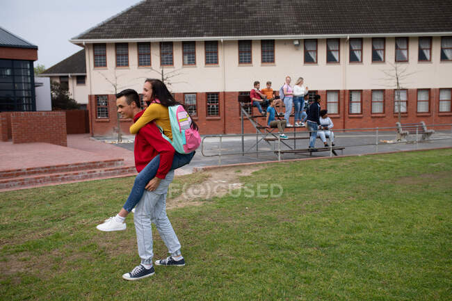 Vista lateral de uma mulher caucasiana e casal estudante do ensino médio pendurado para fora, de pé em seu campo de jogo escola, o menino piggybacking a menina, com colegas de classe pendurados e falando no fundo — Fotografia de Stock