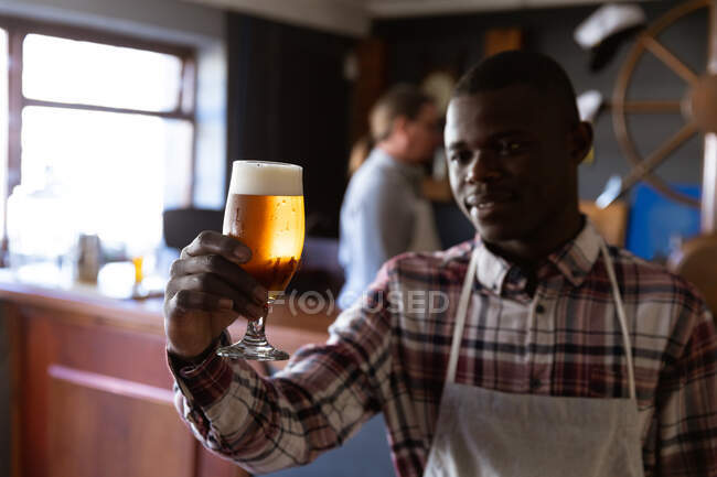 Hombre afroamericano trabajando en un pub de microcervecería, vistiendo delantal blanco, inspeccionando una pinta de cerveza, sosteniéndola frente a él. - foto de stock