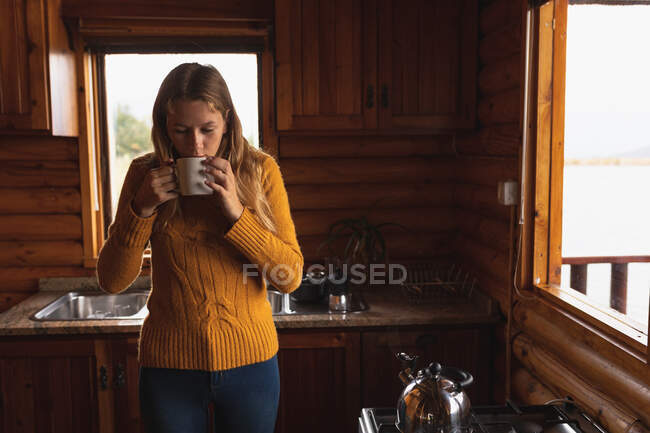 Front view of a Caucasian woman having a good time on a trip to the mountains, standing in a wooden cabin, drinking coffee — Stock Photo