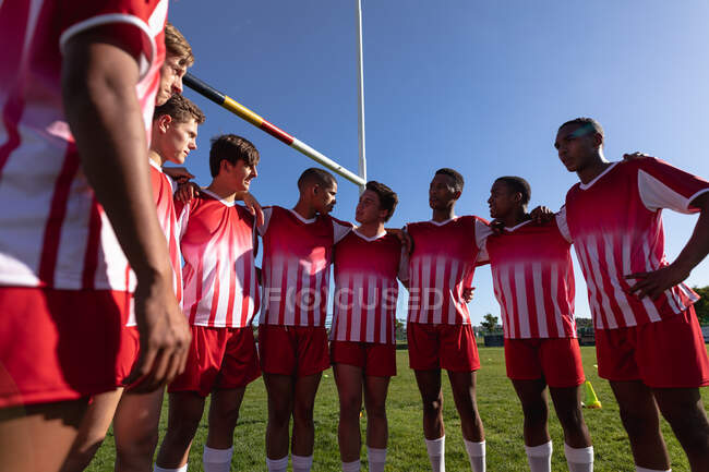 Vista frontal de un equipo masculino multiétnico adolescente de jugadores de rugby que usan su tira de equipo y están parados en el campo de juego en un semicírculo con los brazos alrededor unos de otros, hablando - foto de stock