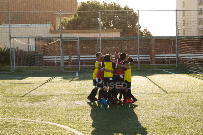 Vista lateral de un grupo multiétnico de jugadores de fútbol de niño de pie en un grupo en un campo de juego en el sol, abrazándose y celebrando ganar el partido - foto de stock
