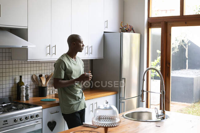 Seitenansicht eines afroamerikanischen Mannes zu Hause, der in der Küche mit einer Tasse Kaffee steht und aus dem Fenster in den Garten blickt — Stockfoto
