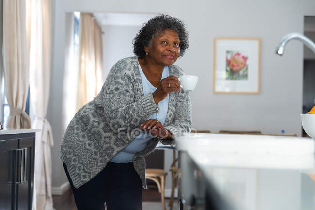 Mulher de raça mista sênior desfrutando de seu tempo em casa, distanciamento social e auto-isolamento em quarentena, de pé em sua cozinha, segurando uma xícara e sorrindo — Fotografia de Stock