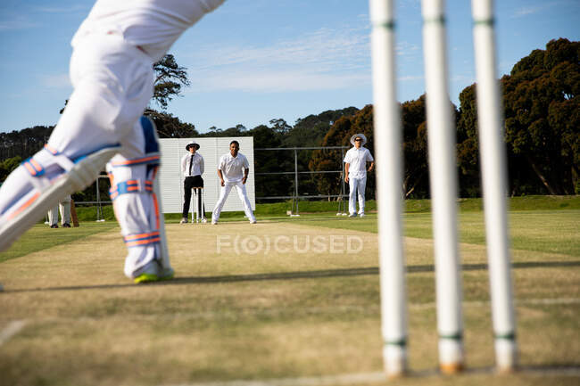 Vue arrière section basse d'un adolescent joueur de cricket blanc sur le terrain lors d'un match de cricket, avec d'autres joueurs jouant en arrière-plan. — Photo de stock