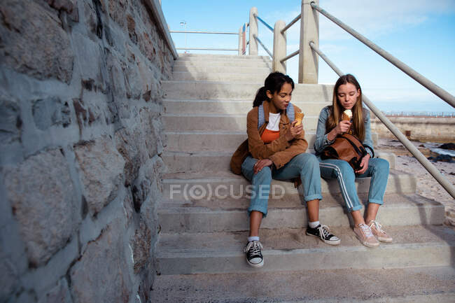 Передній вид на кавказьку і змішану расу дівчат, які відпочивають разом у сонячний день, їдять морозиво, сидячи на сходах під морем.. — стокове фото