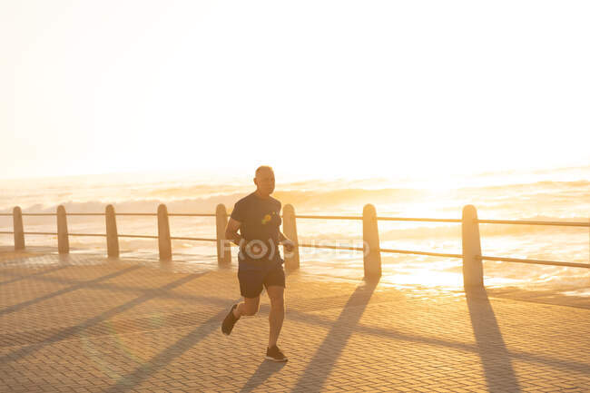 Vista frontal de un anciano caucásico maduro haciendo ejercicio en un paseo marítimo en un día soleado con cielo azul, corriendo al atardecer - foto de stock