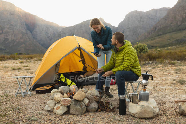 Seitenansicht eines kaukasischen Paares, das sich bei einem Ausflug in die Berge amüsiert: Ein Mann sitzt am Lagerfeuer und kocht Würstchen auf den Stöcken, während eine Frau steht und ihm etwas auf einem Smartphone zeigt. — Stockfoto