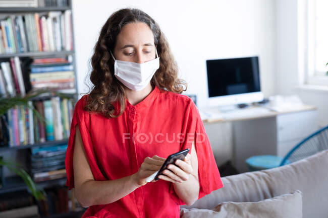 Caucasian woman spending time at home, wearing a pink dress and a face mask against coronavirus, covid 19, cleaning her smartphone. Social distancing and self isolation in quarantine lockdown. — Stock Photo