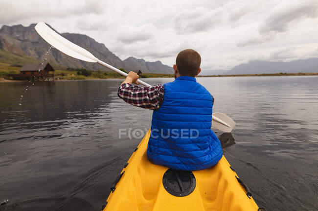 Rückansicht eines kaukasischen Mannes, der sich bei einem Ausflug in die Berge amüsiert, auf einem See Kajak fährt und die Aussicht genießt — Stockfoto