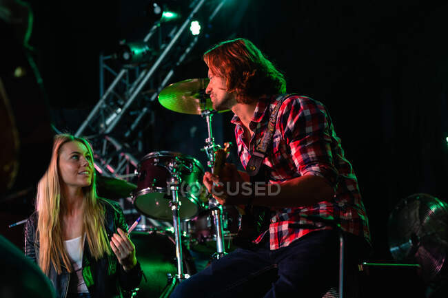 Side view of a Caucasian male guitarist and a Caucasian female singer rehearsing at a music venue before a performance, sitting and looking at each other while they run through a song together on the stage — Stock Photo