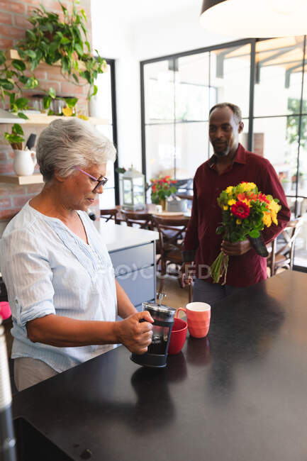 Um casal afro-americano sênior desfrutando de sua aposentadoria, de pé em sua cozinha em um dia ensolarado, a mulher fazendo café e o homem segurando um buquê de flores, e sorrindo antes de dá-los a sua esposa — Fotografia de Stock