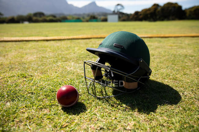 Крупный план красного мяча для крикета и зеленого шлема для крикета, лежащего на поле для крикета в солнечный день — стоковое фото