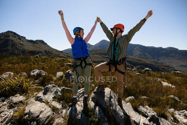 Vue de face du couple caucasien profitant du temps dans la nature, portant un équipement de tyrolienne, se tenant les mains les bras en l'air, se souriant par une journée ensoleillée dans les montagnes — Photo de stock