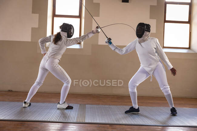 Caucasian and African American sportswomen wearing protective fencing outfit during a fencing training session, taking aim at each other with their epees. Fencers training at gym. — Stock Photo