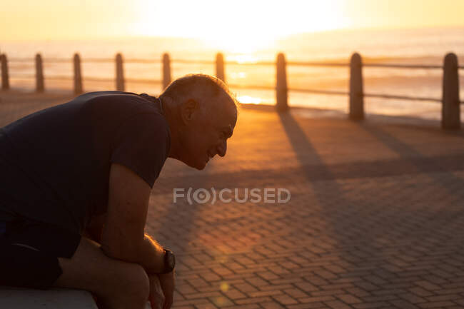 Вид сбоку на взрослого кавказца, тренирующегося на набережной в солнечный день, отдыхающего, сидящего на закате — стоковое фото