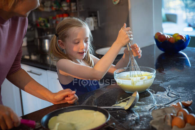 Vista lateral de una mujer caucásica disfrutando de tiempo en familia con su hija en casa juntos, cocinando, haciendo panqueques usando batidor de huevos y sonriendo en su cocina - foto de stock
