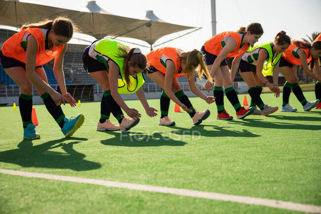 Вид сбоку на группу кавказских хоккеистов, тренирующихся перед игрой, тренирующихся на поле для хоккея на траве, делающих упражнения, растягивающих ноги, наклоняющихся к ногам в солнечный день — стоковое фото