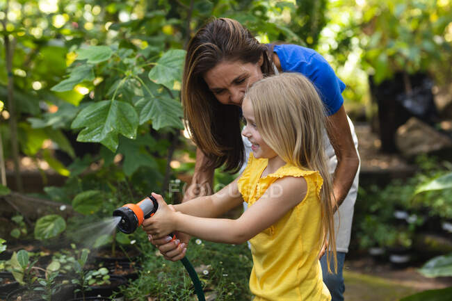 Une femme caucasienne et sa fille s'amusent ensemble dans un jardin ensoleillé, en utilisant un tuyau d'arrosage pour arroser les plantes — Photo de stock