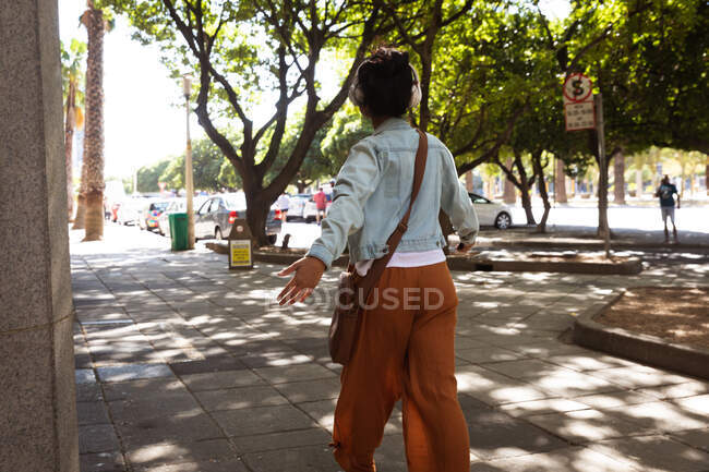 Rückansicht einer Mischlingsfrau mit langen dunklen Haaren, die tagsüber auf den Straßen der Stadt unterwegs ist, eine Jeansjacke trägt und in einer Stadtstraße mit Bäumen und Autos im Hintergrund spaziert. — Stockfoto