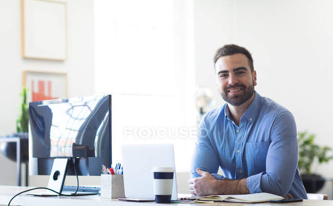 Retrato de un hombre de negocios caucásico con el pelo corto, con una camisa azul, trabajando en una oficina moderna, sentado en una mesa y sonriendo, mirando a la cámara - foto de stock