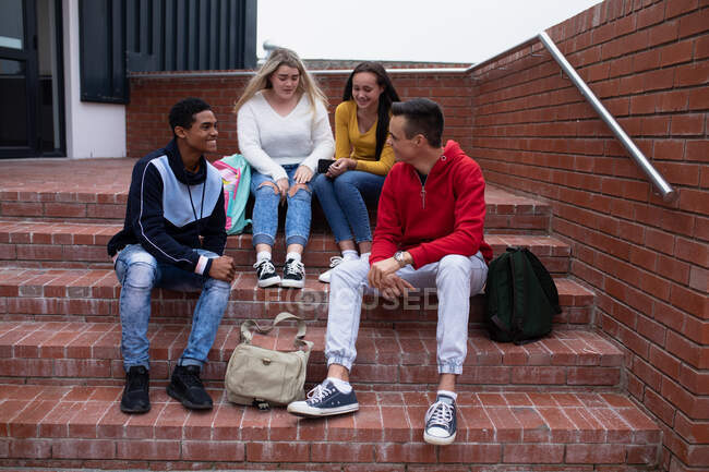 Вид спереди на многоэтническую группу старшеклассников, юношей и девушек со школьными сумками, которые болтаются и разговаривают, сидя на ступеньках на школьной территории — стоковое фото