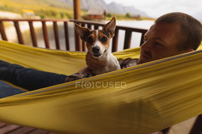 Vista lateral de cerca de un hombre caucásico pasar un buen rato en un viaje a las montañas, acostado en una hamaca con un cachorro en el pecho - foto de stock