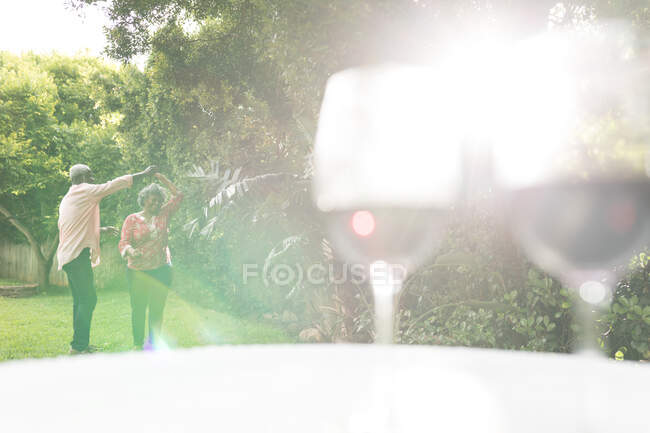 Un couple afro-américain âgé passe du temps dans leur jardin ensemble, la distance sociale et l'isolement personnel en quarantaine pendant l'épidémie de coronavirus covid 19, dansant avec des verres de vin rouge au premier plan — Photo de stock