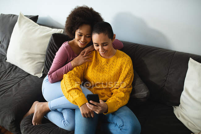 Vista frontal do casal feminino misto relaxando em casa, sentado em um sofá abraçando e olhando para um smartphone — Fotografia de Stock
