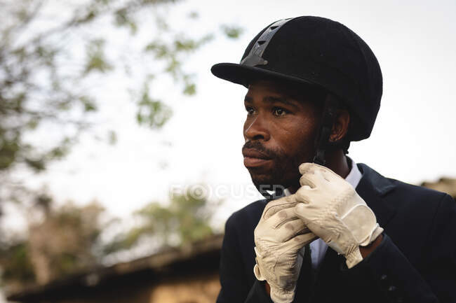Side view close up of a smartly dressed African American man putting a riding hat on before dressage horse riding during a sunny day. — Stock Photo